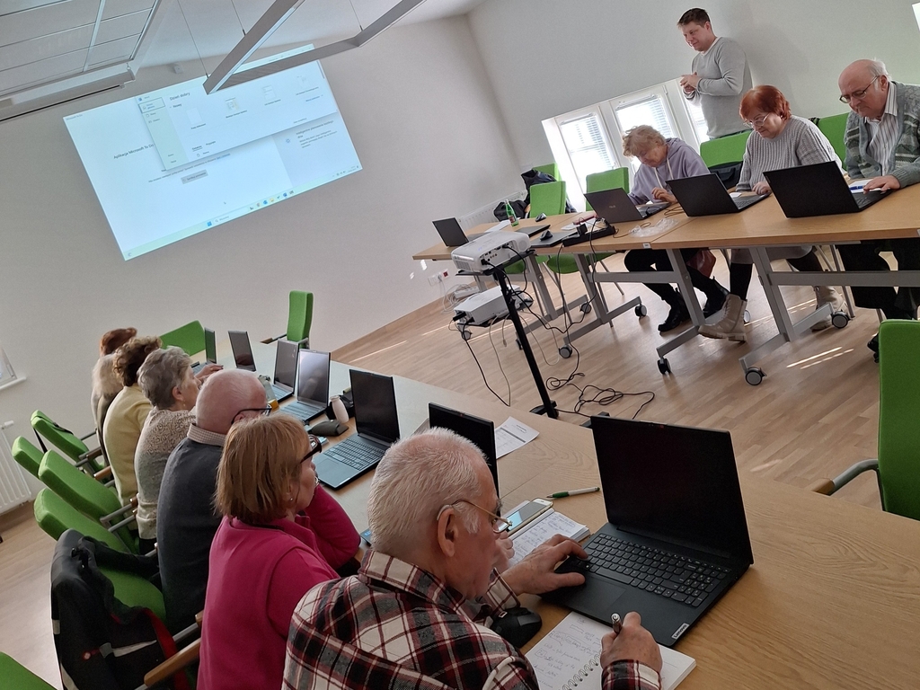 Zajęcia medialne. Kilkunastu seniorów siedzi przy stolikach w dwóch rzędach, zwróconych przodem do siebie. Mają otwarte, ale nieuruchomione laptopy. Mężczyzna stojący w końcu sali pod oknem cos mówi. Z lewej strony na ścianie wyświetla się slajd.