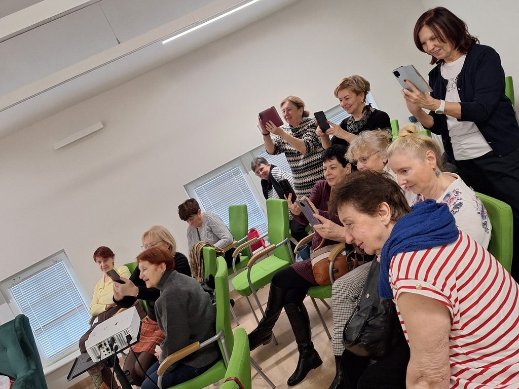 Spotkanie z podróżniczką Zofia Suską. Seniorki słuchają z zaangażowaniem. Niektóre siedzą, inne stoją. Kilka robi zdjęcia. 
