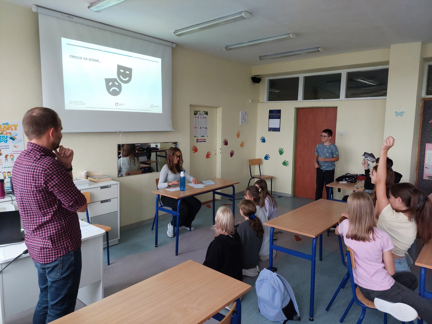 Dzieci w klasie zgłaszają się do odpowiedzi, na środku klasy siedzi dziewczynka odgrywająca scenkę teatralną
