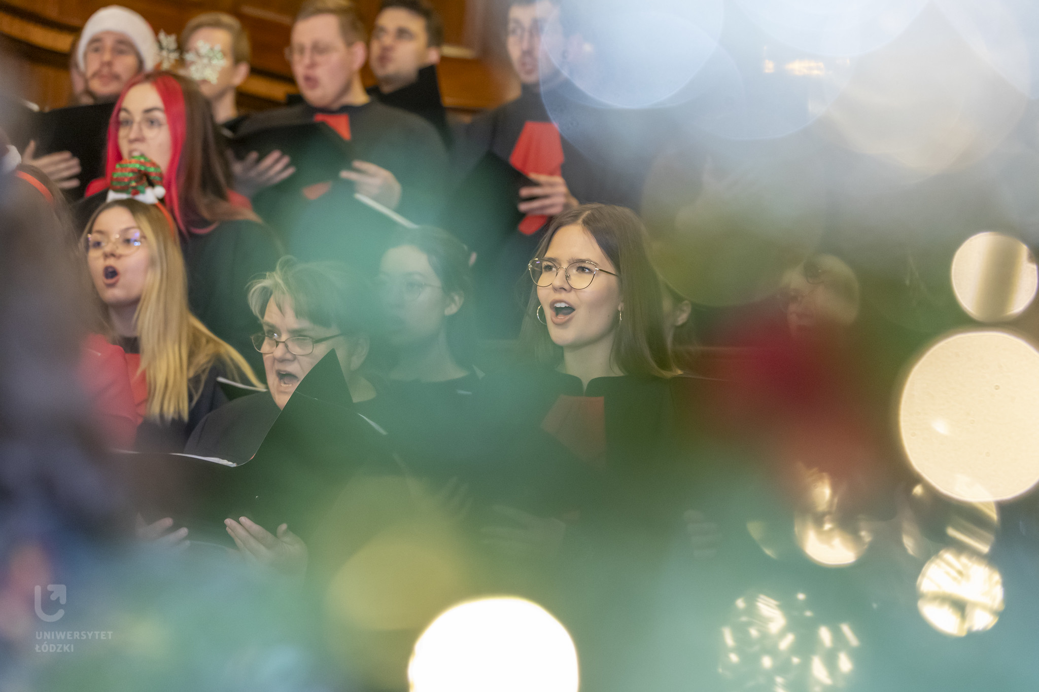Na zdjęciu widać chór ludzi w trakcie śpiewu. Na pierwszym planie widoczne są rozmyte światła lampek choinkowych i fragmenty gałązek. 