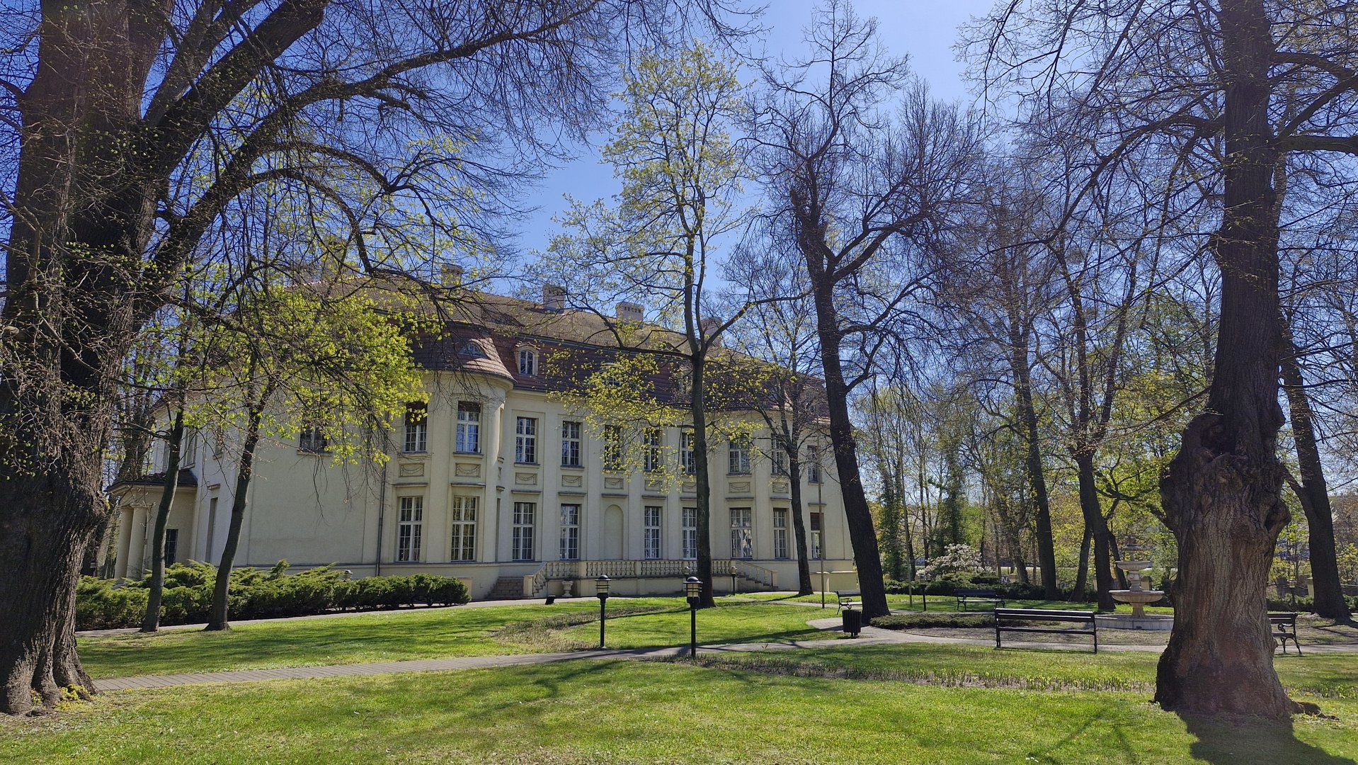 Na zdjęciu widać fragment parku, drzewa, trawę, fontannę oraz w oddali duży budynek - pałac.