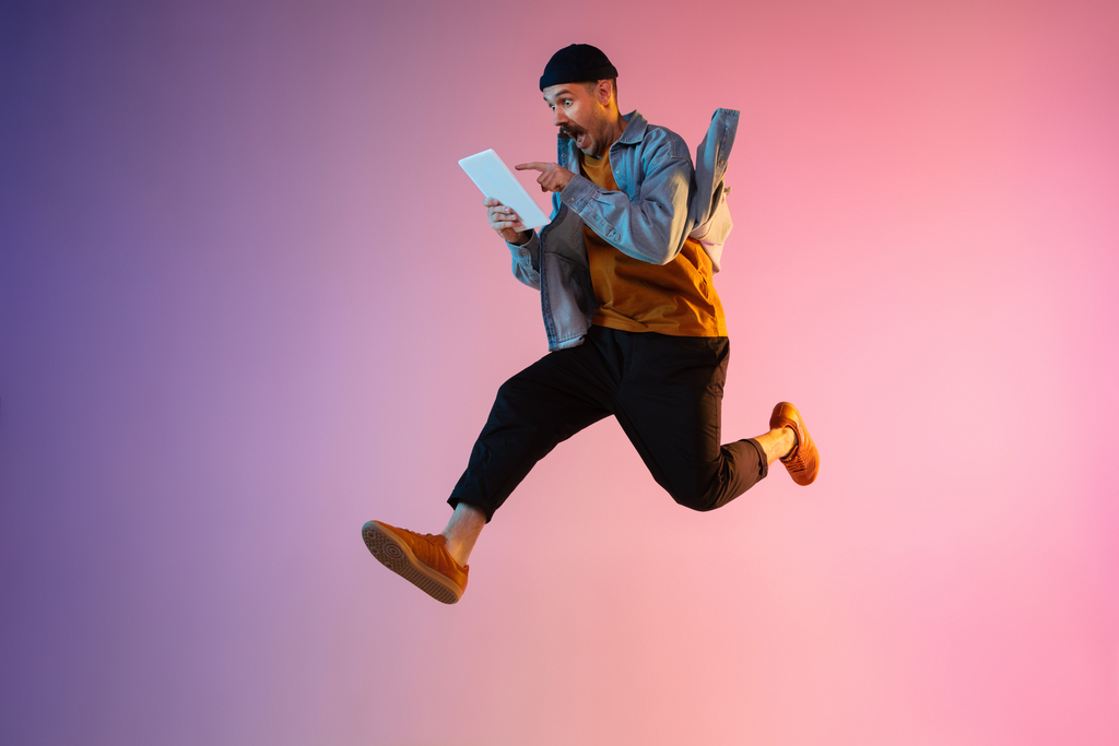 Full length portrait of happy jumping man wearing casual clothes in neon light isolated on gradient background. Emotions, ad concept. Using tablet, hurrying up, late for work or sale, shopping. Grafika rekrutująca