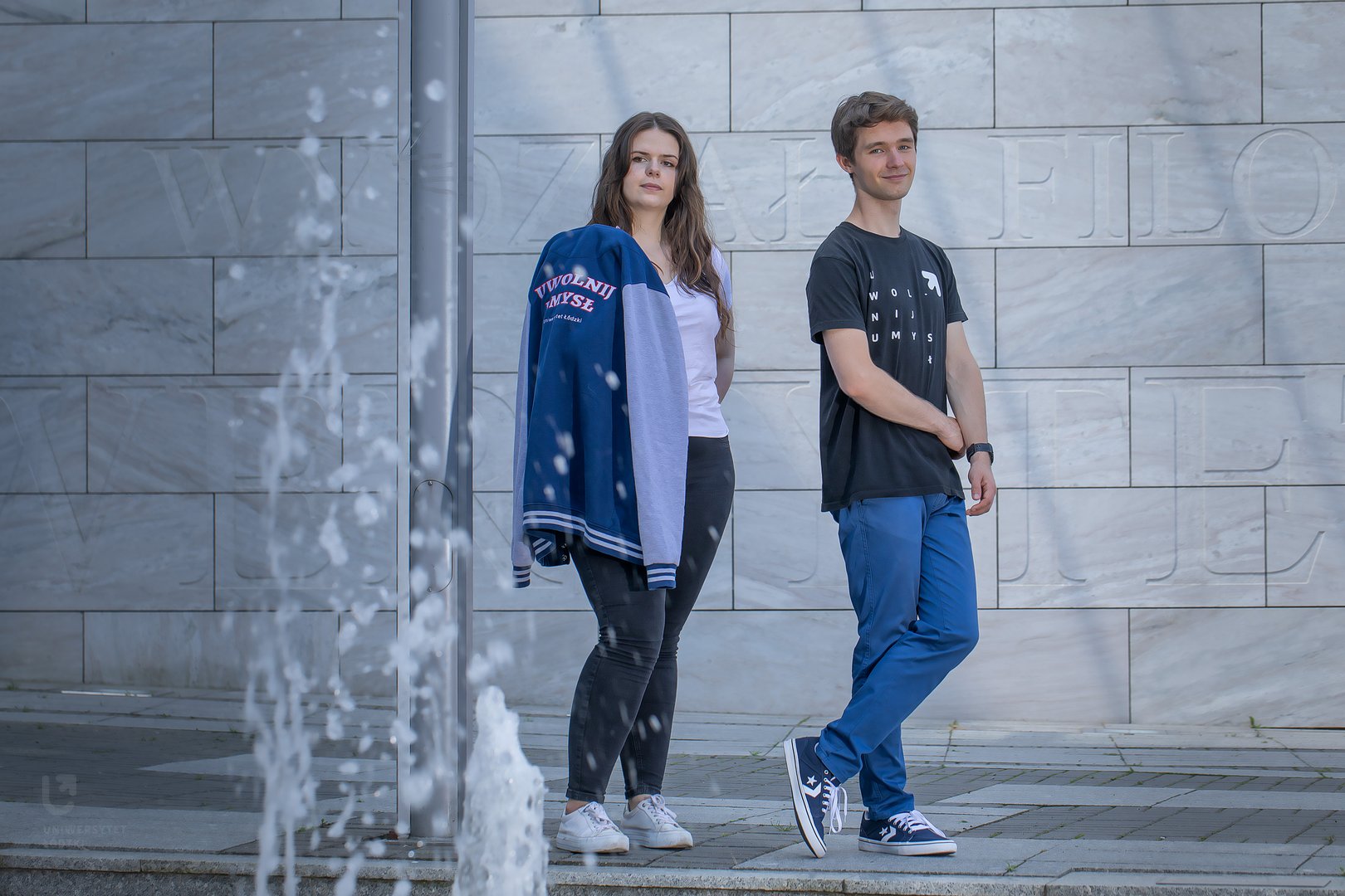 Student i studentka na tle szarej ściany. Przed nimi, ma pierwszym planie, tryska woda z fontanny