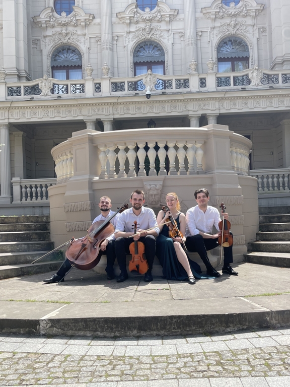 Kobieta i 3 mężczyzn - członkowie zespołu Vivenda String Quartet, siedzą z instrumentami smyczkowymi przed budynkiem