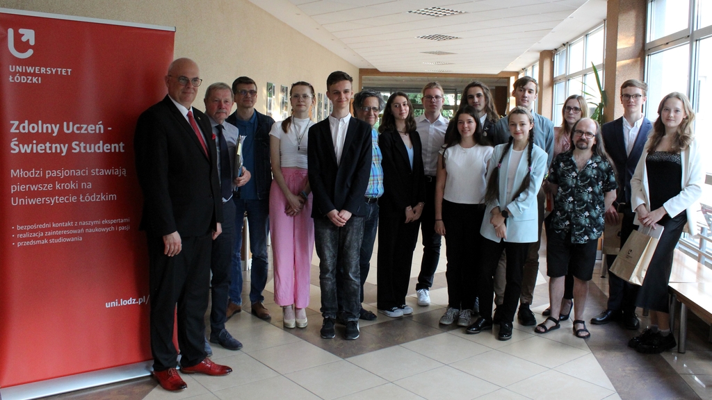 Grupa młodych i starszych osób stojących w korytarzu budynku. Wszyscy są elegancko ubrani, niektórzy z nich trzymają dokumenty. Po lewej stronie widoczny jest czerwony roll-up z napisem "Zdolny Uczeń - Świetny Student" . W tle widać okna, przez które wpada naturalne światło.