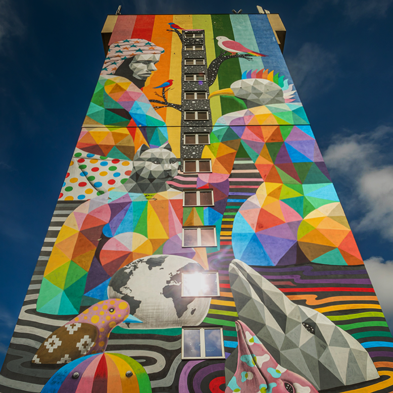 Na zdjęciu znajduje się wielobarwny mural pokrywający całą fasadę wysokiego budynku. Obraz składa się z geometrycznych, wielościennych form tworzących postacie ludzi i zwierząt — widoczne są m.in. delfin, ptaki, niedźwiedź i głowa człowieka w profilu. Elementy są pomalowane w żywe, kontrastowe kolory tworzące mozaikę w stylu low poly. Centralnie przez mural biegną okna w pionowym rzędzie, a w tle widoczne jest niebieskie niebo z lekkim zachmurzeniem. Mural przedstawia różnorodność biologiczną i jedność ludzi z naturą, tworząc symboliczny i optymistyczny przekaz.